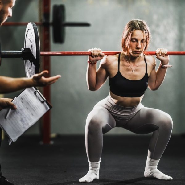 Muscular young woman working out with personal trainer at the gym. She is doing squat exercise with barbell.